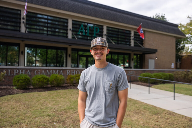 Wyatt Harvey smiling and standing in front of a building with the letters "ΑΓΡ" displayed. The building has a sign reading "Alpha Gamma Rho" and features flags above.
