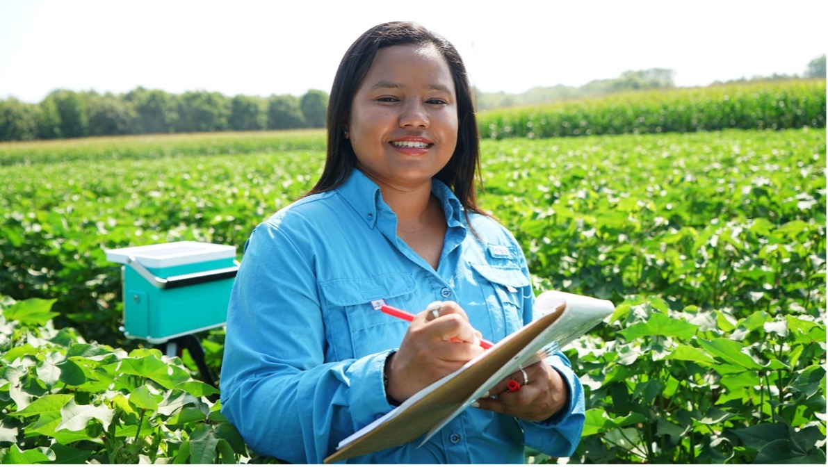 Smitri Shahi in a blue shirt stands in a lush green field, holding a clipboard and pen, with a bright smile.