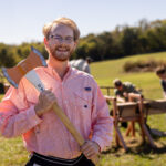 Max Brabson stands smiling while holding an axe in a grassy field with a small group of people working with wood in the background. A tree line is visible in the distance under a clear blue sky.