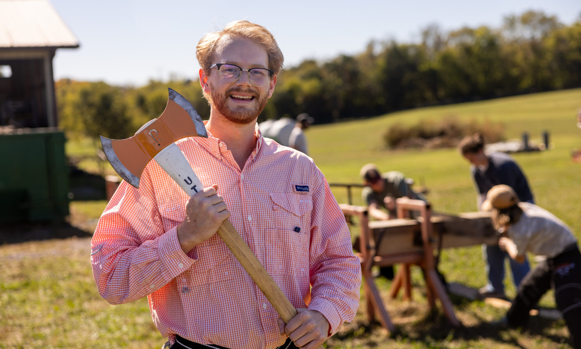 Max Brabson stands smiling while holding an axe in a grassy field with a small group of people working with wood in the background. A tree line is visible in the distance under a clear blue sky.
