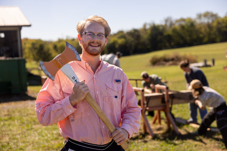 Max Brabson stands smiling while holding an axe in a grassy field with a small group of people working with wood in the background. A tree line is visible in the distance under a clear blue sky.