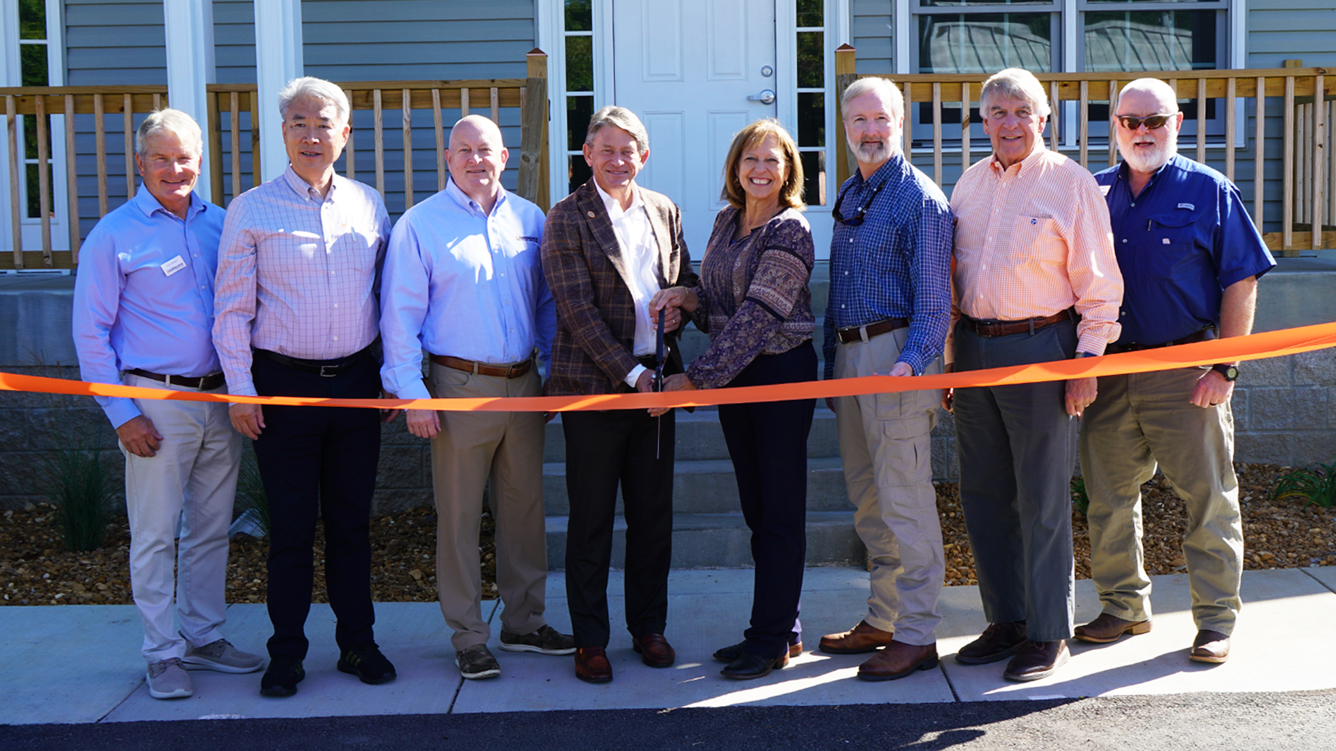 Eight people, two holding a pair of scissors, pose for a picture behind an orange ribbon at the front of a house